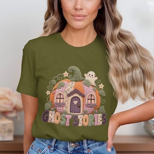 Op de afbeelding: Een olijfgroen T-shirt met korte mouwen en een afbeelding van een pompoenvormig huis met een paarse deur, ramen en een klein spookje. Het ontwerp bevat een patchworkpatroon in oranje, roze en groen, met de tekst GHOST STORIES.