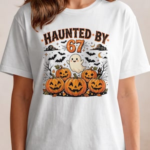 Op de afbeelding: Een wit t-shirt met een grafische print met de tekst HAUNTED BY 67, een kleine witte geest, vier oranje pompoenen, zwarte vleermuizen, spinnenwebben, sterren en een halve maan in een feestelijke Halloween-illustratie.