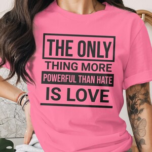 Op de afbeelding: Een felroze t-shirt met de tekst "THE ONLY THING MORE POWERFUL THAN HATE IS LOVE" in zwarte letters. De tekst is in drie regels binnen een rechthoekig kader geplaatst.