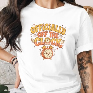 Op de afbeelding: Een wit T-shirt met korte mouwen en een grafische print in oranje en gele tinten. De tekst "OFFICIALLY OFF THE CLOCK" staat boven een illustratie van een wekker met een lachend gezicht, kleine vleugels en decoratieve steraccenten.
