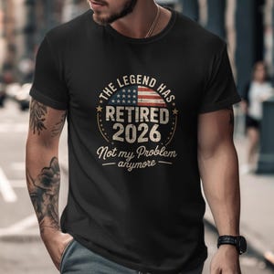 May include: Black t-shirt with a circular graphic featuring the American flag and the text "THE LEGEND HAS RETIRED 2026 Not my Problem anymore". The shirt is worn by a person.