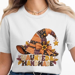 Op de afbeelding: Een wit t-shirt met een afbeelding van een heksenhoed gemaakt van oranje en zwart geruite, bloemrijke en effen stoffen lappen. De hoed is versierd met een zonnebloem, bladeren en een hangende ster. De tekst HAUNTED & HANDMADE staat eronder in blokletters.
