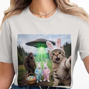 May include: A light gray t-shirt featuring a humorous graphic. The graphic depicts a cat wearing bunny ears taking a selfie with a UFO, Bigfoot, and two bunnies in suits. A basket of Easter eggs is also present.