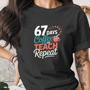 Op de afbeelding: Zwart T-shirt met de tekst "67 Days of Coffee Teach Repeat" in wit, teal, rood en crème kleuren. Het ontwerp bevat een koffiekopje. Het shirt is een casual kledingstuk.