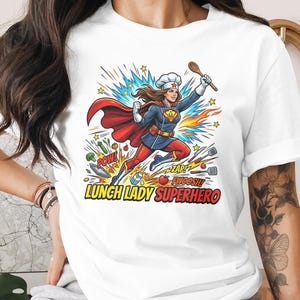 Op de afbeelding: Wit t-shirt met een cartoon illustratie van een superheld kok. Het ontwerp toont een kok met een cape, die een houten lepel vasthoudt, met de tekst "LUNCH LADY SUPERHERO" eronder. Kleurrijke actiewoorden zoals "POW!" en "ZAP!" zijn ook inbegrepen.