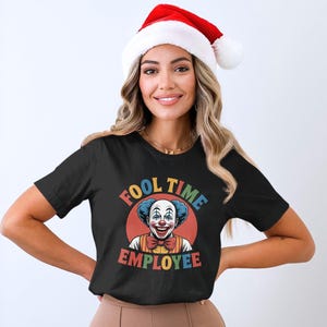 Op de afbeelding: Zwart T-shirt met een clownafbeelding en de tekst "FOOL TIME EMPLOYEE". De clown heeft blauw haar, een rode neus en een vlinderdas. De persoon draagt een rood-witte kerstmuts.
