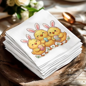 Op de afbeelding: Een stapel witte vierkante servetten met een paasillustratie. Het ontwerp toont drie gele kuikens met konijnenoren die kleurrijke paaseieren vasthouden. De servetten zijn gestapeld op een houten oppervlak.