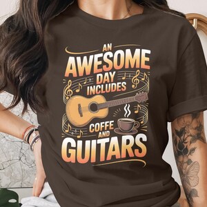 Op de afbeelding: Bruin t-shirt met een grafisch ontwerp met een gitaar, koffiekopje en muzieknoten. De tekst luidt "An Awesome Day Includes Coffee and Guitars" in oranje en wit. Een casual kledingstuk.