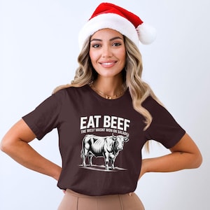Op de afbeelding: Een donkerbruin t-shirt met de tekst "EAT BEEF" boven een zwart-wit illustratie van een stier. De tekst "THE WEST WASN'T WON ON SALADS" staat eronder. De persoon draagt een rood-witte kerstmuts.