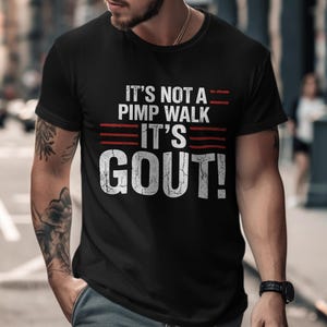 Op de afbeelding: Zwart T-shirt met de tekst "IT'S NOT A PIMP WALK IT'S GOUT!" in verweerde witte letters, met rode horizontale lijnen. Het T-shirt wordt gedragen door een persoon met tatoeages op zijn arm.