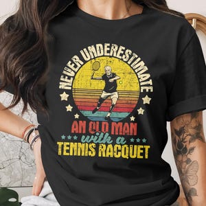 Op de afbeelding: Zwart T-shirt met een vintage ontwerp met een tennisspeler en de tekst "NEVER UNDERESTIMATE AN OLD MAN WITH A TENNIS RACQUET". Het ontwerp bevat een zonsondergang achtergrond en ster accenten.