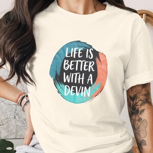 Può includere: T-shirt color crema con la scritta bianca "LIFE IS BETTER WITH A DEVIN", inserita in un design circolare blu, nero e corallo. La maglietta ha le maniche arrotolate.