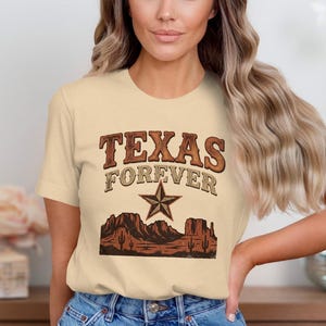 Op de afbeelding: Een crèmekleurig T-shirt met korte mouwen en een grafische print met de tekst "TEXAS FOREVER" in bruine letters boven een bruine ster en een illustratie van een woestijnlandschap met mesa's en cactussen in bruine en donkeroranje tinten.
