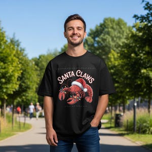 Op de afbeelding: Zwart T-shirt met een feestelijke afbeelding van een rode kreeft met een kerstmuts en kerstverlichting. De tekst "Santa Claws" staat boven de kreeft. Het shirt wordt buiten door een persoon gedragen.
