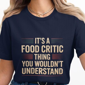 Op de afbeelding: Marineblauw T-shirt met de tekst "IT'S A FOOD CRITIC THING YOU WOULDN'T UNDERSTAND" in een retro lettertype. De tekst is beige met horizontale strepen erboven en eronder. Het shirt heeft een ronde hals.