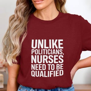 Op de afbeelding: Een bordeauxrood T-shirt met de witte tekst: "UNLIKE POLITICIANS, NURSES NEED TO BE QUALIFIED". Het shirt is gemaakt van een zacht materiaal en is geschikt voor casual wear.