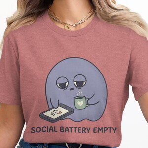 Pode incluir: Uma t-shirt de manga curta em rosa seco com um gráfico de um fantasma roxo cansado a segurar uma caneca verde com um coração e a olhar para um smartphone com um ícone de bateria a 1%. O texto abaixo do gráfico diz SOCIAL BATTERY EMPTY.