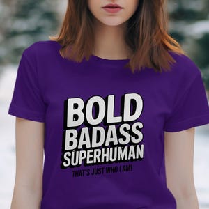 Può includere: Una maglietta viola con la scritta "BOLD BADASS SUPERHUMAN" in bianco, bordata di nero. Sotto, la frase "THAT'S JUST WHO I AM!". La maglietta ha maniche corte e scollo tondo.