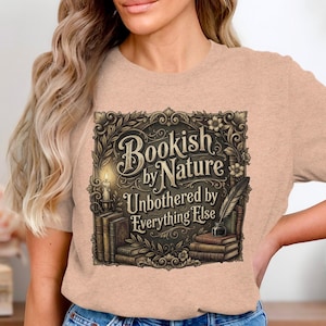 Op de afbeelding: Een lichtbruin t-shirt met korte mouwen en een donkerbruine grafische print van boeken, een brandende kaars en een ganzenveer. De tekst luidt Bookish by Nature Unbothered by Everything Else, omringd door een sierlijke, bloemrijke rand.