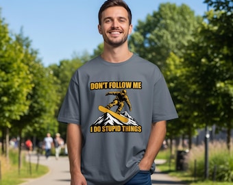 Könnte beinhalten: Graues T-Shirt mit dem Text "DON'T FOLLOW ME I DO STUPID THINGS" und einer gelb-schwarzen Grafik eines Snowboarders. Das T-Shirt wird von einer Person getragen, mit einem Hintergrund aus Bäumen und einem Weg.