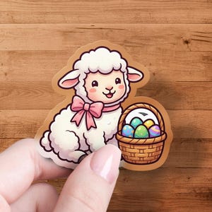 Op de afbeelding: Een sticker met een cartoon lammetje met een roze strik, naast een mand met kleurrijke paaseieren. Het lammetje is wit met een lachend gezicht. De mand is bruin en de eieren zijn versierd.