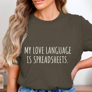 May include: Olive green t-shirt with the text "MY LOVE LANGUAGE IS SPREADSHEETS" in white. The shirt is a crew neck style and is worn by a person with long blonde hair and blue jeans.
