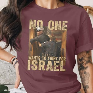 May include: Burgundy t-shirt featuring a distressed graphic with the text "NO ONE wants to fight for ISRAEL." The graphic depicts two figures in uniform with an American flag in the background. The shirt has short sleeves.