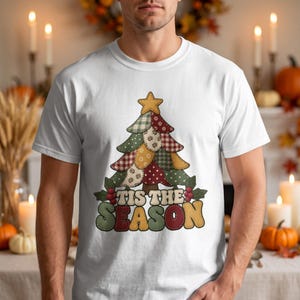 Op de afbeelding: Een wit T-shirt met korte mouwen en een afbeelding van een kerstboom in patchworkstijl, bekroond met een gele ster. Onder de boom staat de tekst "TIS THE SEASON" in grote, veelkleurige, afgeronde blokletters met hulstaccenten.