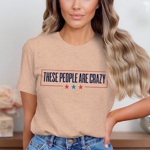 Op de afbeelding: Lichtperzikkleurig T-shirt met de tekst "THESE PEOPLE ARE CRAZY" in een marineblauwe rechthoek met rode en blauwe sterren. Het shirt wordt gedragen door een persoon met lang, golvend blond haar.