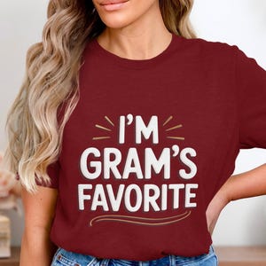 Op de afbeelding: Een bordeauxrood t-shirt met de tekst "I'M GRAM'S FAVORITE" in wit, omlijnd met een donkerdere kleur. De tekst staat in het midden van het shirt, met decoratieve lijnen erboven en eronder. Het shirt is effen van kleur.