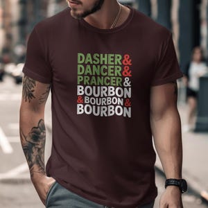 Op de afbeelding: Bordeauxrood t-shirt met de tekst "DASHER & DANCER & PRANCER & BOURBON & BOURBON & BOURBON" in groen, rood en wit. Het shirt heeft een ronde hals en korte mouwen.