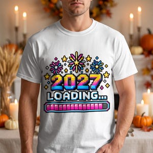 May include: A white short-sleeve t-shirt featuring a pixel art graphic. The design displays the text 2027 LOADING... in a colorful gradient font, accompanied by pixelated fireworks and a pink progress bar at the bottom.