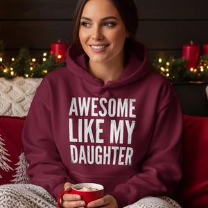 Op de afbeelding: Bordeaux hoodie met de tekst "AWESOME LIKE MY DAUGHTER" in het wit. De hoodie heeft een capuchon met trekkoord en een voorzak. De persoon houdt een rode mok vast. De achtergrond bevat kerstversieringen.