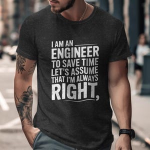 Op de afbeelding: Donkergrijs t-shirt met de tekst "I AM AN ENGINEER TO SAVE TIME LET'S ASSUME THAT I'M ALWAYS RIGHT." Het shirt wordt gedragen door een persoon met tatoeages op zijn arm en een horloge om zijn pols.