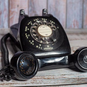 May include: A black rotary dial phone with a black cord and a black handset. The phone has a white dial with black numbers and letters. The phone is on a wooden surface.