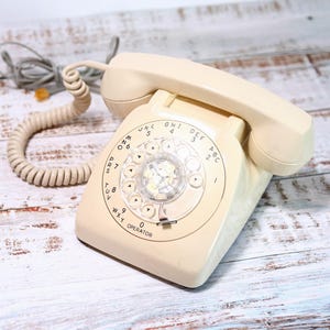 May include: A vintage rotary dial phone with a cream-colored body and a black rotary dial. The phone has a cord and is sitting on a white wooden surface.