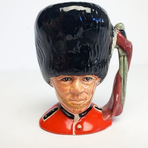 May include: A ceramic mug shaped like a British guardsman's head, wearing a black bearskin hat and red uniform. The mug has a handle shaped like a flag.