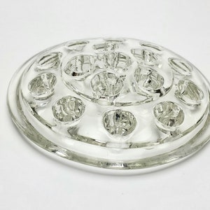May include: A clear glass flower frog with 16 small round openings for holding flower stems. The frog is circular and has a raised rim.