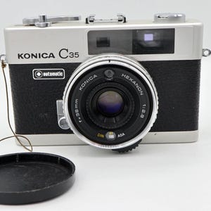 May include: A silver and black Konica C35 automatic camera with a Hexanon 1:2.8 lens. The camera has a black lens cap and the text "KONICA C35" is visible on the front. The lens is marked with "f=38mm".