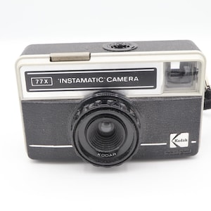 May include: A vintage Kodak Instamatic 77X camera. The camera is black with a silver top and a prominent lens. The text "77X 'INSTAMATIC' CAMERA" is displayed on the front. A black strap is attached to the side. Made in England.