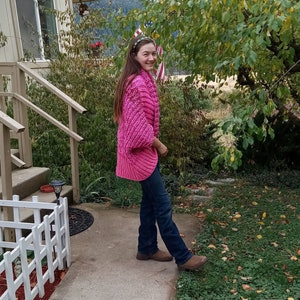 May include: A woman wearing a pink knitted cardigan with a blue jean and brown boots. She is standing on a concrete walkway in front of a house with a white picket fence.