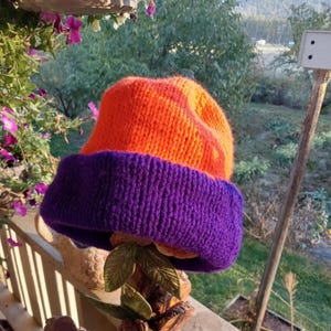 May include: A hand-knitted beanie hat in vibrant orange and purple. The hat features a folded brim in a deep purple color, contrasting with the bright orange crown. The hat is displayed on a decorative wooden stand, showcasing its handmade quality.