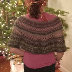 May include: A woman wearing a gray and pink knitted shawl with a pink shirt and blue jeans. The shawl has a lacy pattern and is draped over her shoulders.