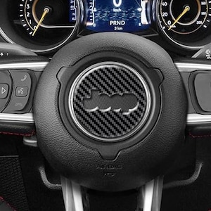 May include: A black leather steering wheel with perforated side grips, red contrast stitching, and a red center stripe at the top. The center hub features a carbon fiber textured Jeep logo emblem, surrounded by silver-toned control buttons and spokes.