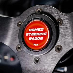 May include: A circular red domed steering wheel badge featuring the white text DOMED STEERING BADGE and a white horn icon. The badge is mounted in the center of a brushed metal steering wheel secured by six visible black hex bolts.