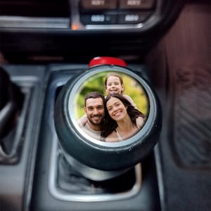 May include: Black gear shift knob with a circular photo of a smiling family. The photo is framed by a silver ring. The gear shift knob is in a car interior.