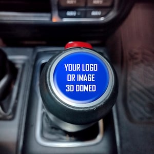 May include: A black gear shift knob with a blue, domed center. The center has the text "YOUR LOGO OR IMAGE 3D DOMED" in white. The knob has a red top and is set in a car's interior.
