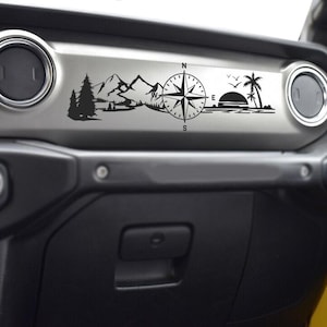 May include: Black vinyl decal featuring a mountain range, compass, and sunset scene. The design includes pine trees, mountains, a compass rose, a setting sun, palm trees, and birds. The decal is designed to be applied to a vehicle's dashboard.
