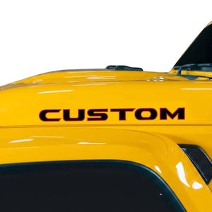 May include: Close-up of a yellow vehicle hood with the word "CUSTOM" in black lettering. The hood has a glossy finish and is partially visible, showing the front of the vehicle.