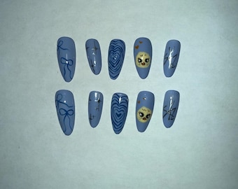 May include: A set of ten light blue, almond-shaped press-on nails. Each nail features a unique design, including bows, hearts, crosses, and a skull with a yellow face. The designs are in shades of blue and silver.